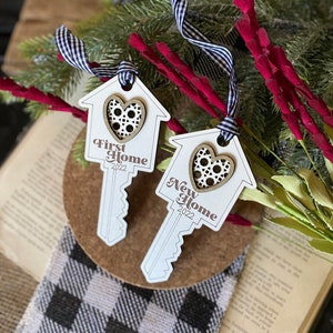 May include: Two wooden key-shaped ornaments with a house design and a heart cutout. The ornaments are white with brown accents and the text "First Home 2022" and "New Home 2022".