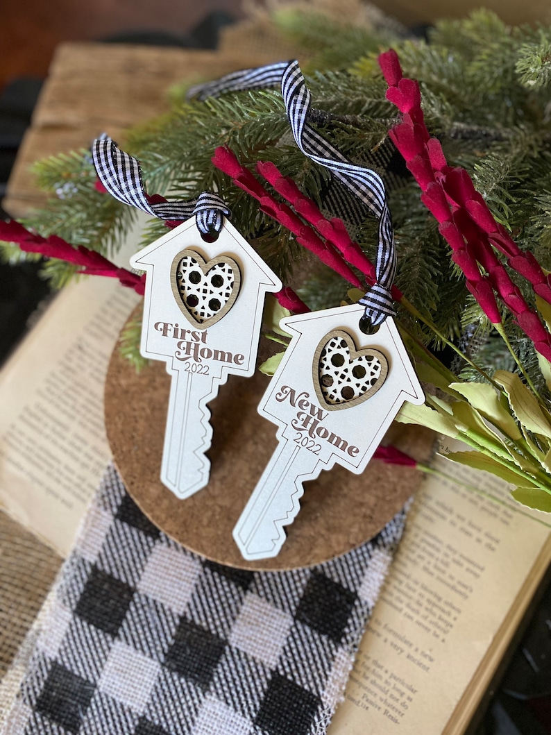 May include: Two wooden key-shaped ornaments with a house design and a heart cutout. The ornaments are white with brown accents and the text "First Home 2022" and "New Home 2022".