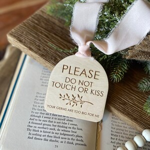 May include: An arched wooden sign with a light beige finish, featuring the engraved text "PLEASE DO NOT TOUCH OR KISS" and "YOUR GERMS ARE TOO BIG FOR ME" with a small leaf illustration. A pale pink ribbon is tied through a hole at the top.