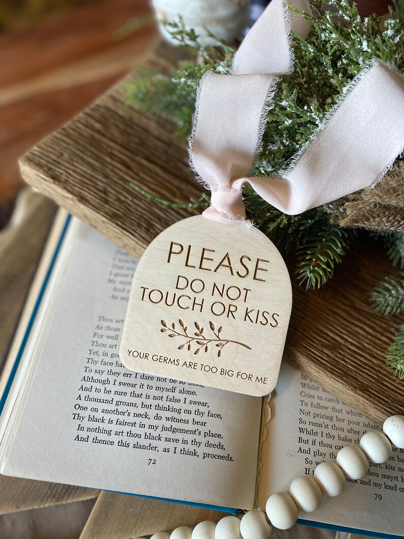 May include: An arched wooden sign with a light beige finish, featuring the engraved text "PLEASE DO NOT TOUCH OR KISS" and "YOUR GERMS ARE TOO BIG FOR ME" with a small leaf illustration. A pale pink ribbon is tied through a hole at the top.