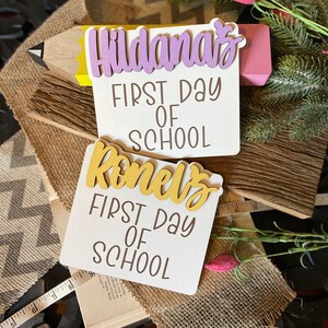 May include: Two white wooden signs with the text "First Day of School" and the names "Hildana's" and "Romel's" in gold and purple letters. The signs are decorated with a pencil and a small sprig of greenery.