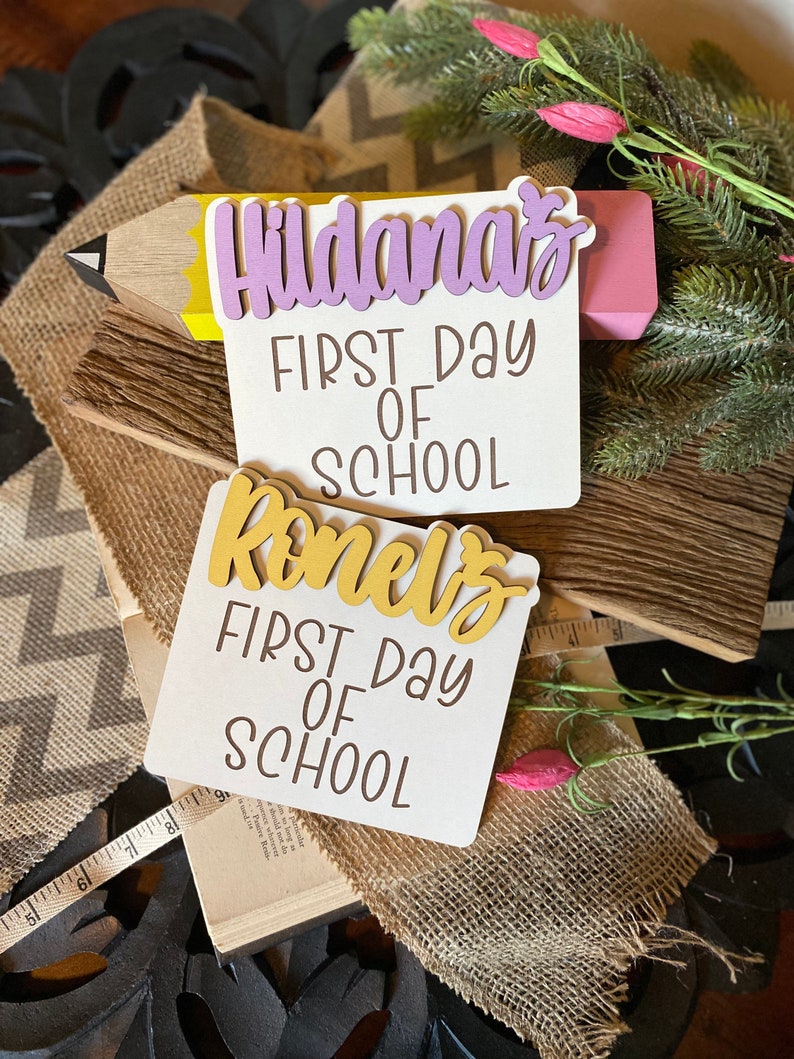 May include: Two white wooden signs with the text "First Day of School" and the names "Hildana's" and "Romel's" in gold and purple letters. The signs are decorated with a pencil and a small sprig of greenery.