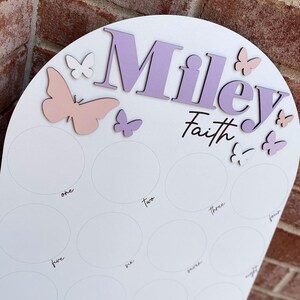 May include: A white wooden arch with the name "Miley Faith" written in purple and pink lettering. The arch is decorated with pink and white butterflies. There are ten circles on the arch, each labeled with a number from one to ten.
