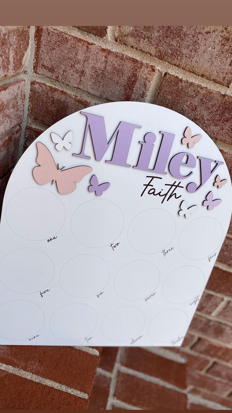 May include: A white wooden arch with the name "Miley Faith" written in purple and pink lettering. The arch is decorated with pink and white butterflies. There are ten circles on the arch, each labeled with a number from one to ten.