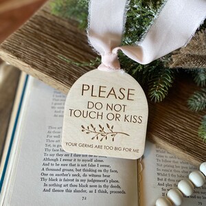 May include: An arched wooden sign with a light tan finish featuring the engraved text PLEASE DO NOT TOUCH OR KISS, a small branch illustration, and the text YOUR GERMS ARE TOO BIG FOR ME, suspended by a pale pink frayed fabric ribbon.
