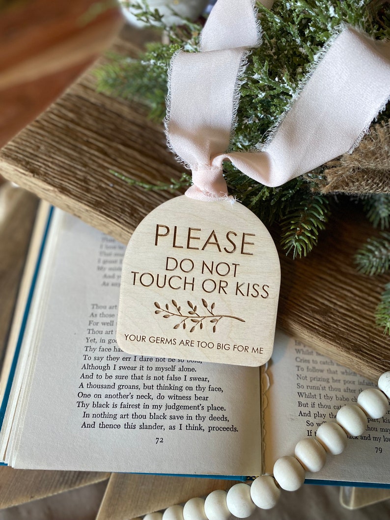 May include: An arched wooden sign with a light tan finish featuring the engraved text PLEASE DO NOT TOUCH OR KISS, a small branch illustration, and the text YOUR GERMS ARE TOO BIG FOR ME, suspended by a pale pink frayed fabric ribbon.
