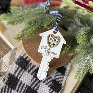 May include: White wooden key-shaped ornament with a house design and a gold heart with a filigree pattern. The ornament says "New Home 2022".