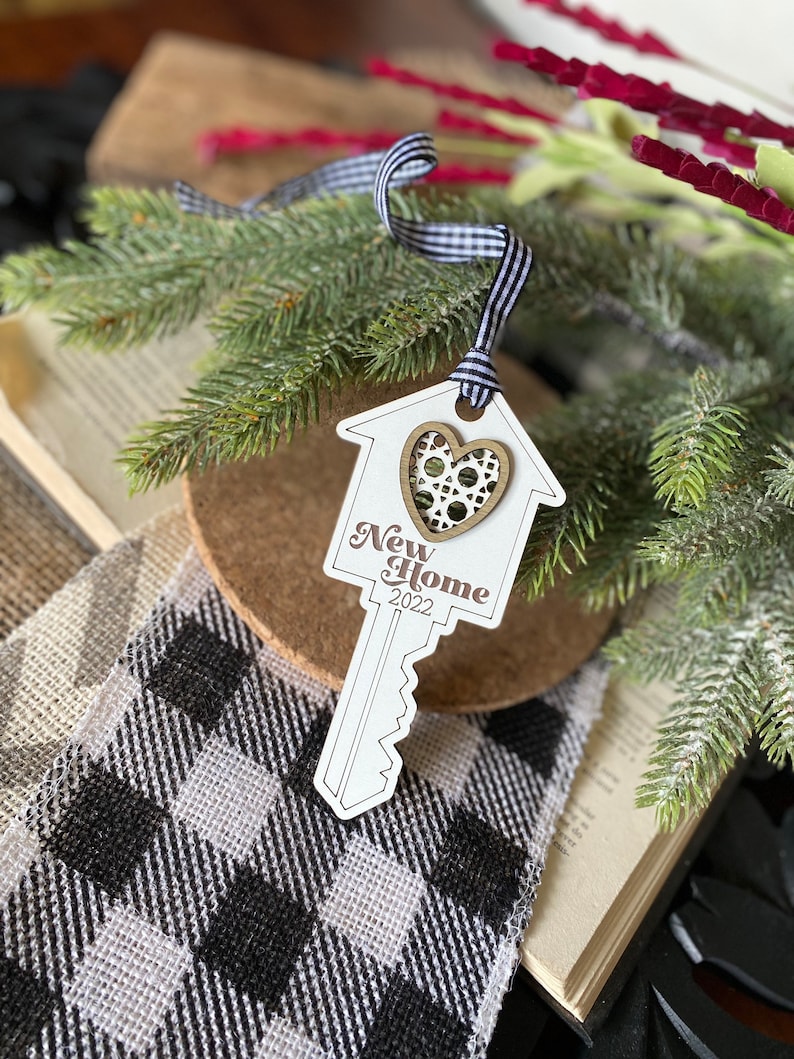 May include: White wooden key-shaped ornament with a house design and a gold heart with a filigree pattern. The ornament says "New Home 2022".