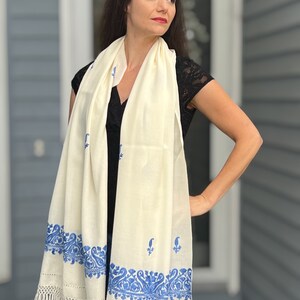 May include: A white shawl with a blue embroidered border. The shawl has a fringe on the bottom edge.