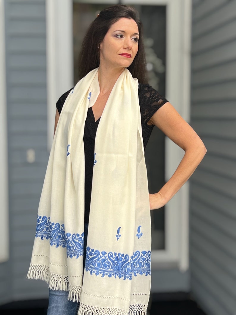 May include: A white shawl with a blue embroidered border. The shawl has a fringe on the bottom edge.