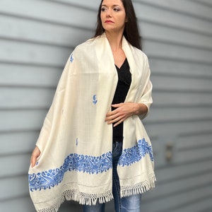 May include: A white shawl with blue embroidery. The shawl has a fringe border and is made of a soft, lightweight fabric.