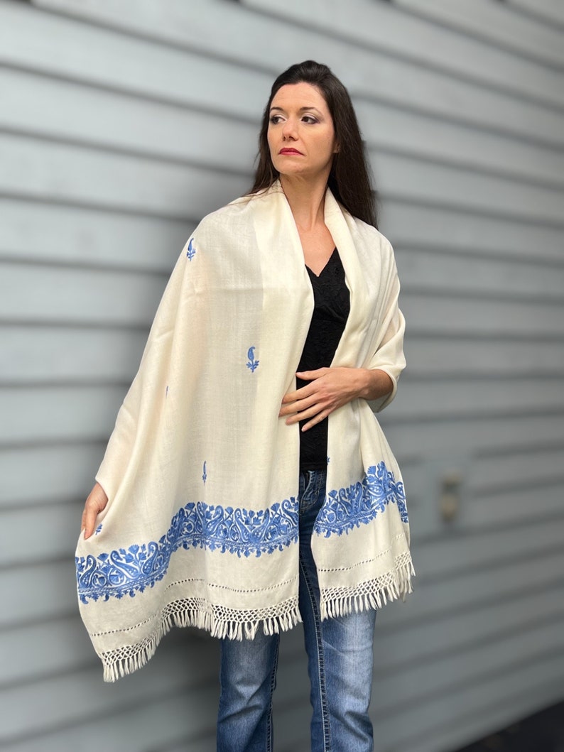 May include: A white shawl with blue embroidery. The shawl has a fringe border and is made of a soft, lightweight fabric.
