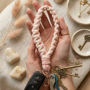 May include: A macrame wristlet keychain featuring a braided pattern in alternating light pink and cream cotton cord. The wristlet is attached to a rose gold metal lobster clasp holding a black car key fob and three assorted metal keys.