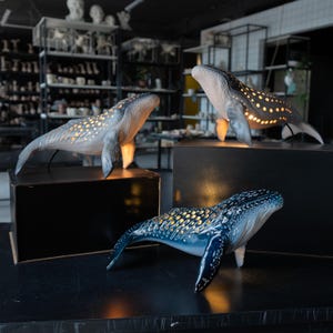 May include: Three whale-shaped lamps with a glossy finish. Two lamps are light gray with illuminated circular cutouts along the back, while the third is a deep blue with similar glowing perforations. Each whale figure is positioned on a black base.