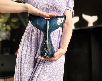 May include: A ceramic whale tail sculpture with a glossy, dark blue finish. The sculpture is held in front of a person wearing a purple floral dress. The tail is a decorative item, possibly for home decor.