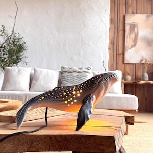 May include: A whale-shaped lamp with a gradient of colours from dark blue to orange, featuring illuminated yellow dots. The lamp is placed on a wooden table, with a black cord extending from its base. The background includes a sofa and decorative elements.