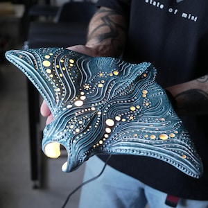 May include: A manta ray shaped lamp with a textured, dark teal surface featuring intricate, swirling patterns and numerous circular cutouts that emit a warm, glowing light from within the hollow, sculpted body of the aquatic creature.