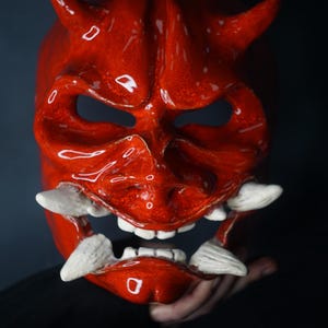 Peut inclure: Un masque Oni rouge brillant avec deux cornes, de grands yeux et une bouche ouverte révélant des crocs blancs. Le masque a une surface texturée et est présenté sur un fond sombre.