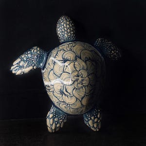 May include: A ceramic turtle figurine with a blue and white floral design. The turtle's shell features a large flower pattern, and its flippers and head are detailed with blue spots. The figurine is set against a dark background.