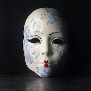 Könnte beinhalten: Eine weiße Keramikmaske mit blauen Koi-Fisch-Designs und roten Akzenten auf Lippen und Wangen. Die Maske hat schwarzes Augen-Make-up und ist vor einem dunklen Hintergrund platziert.