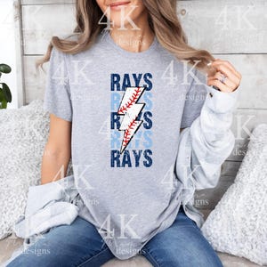 May include: A light gray t-shirt featuring the word "RAYS" in a stacked design with a baseball and lightning bolt graphic. The baseball is white with red stitching, and the lightning bolt is white with a red zig-zag.