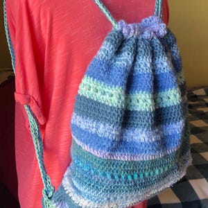 May include: A handmade crochet drawstring backpack in shades of blue, green, and white. The bag features horizontal stripes and a gathered top with a small flower detail. The straps are a light green color.