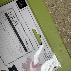 Puede incluir: Un paquete de pegatinas con las palabras "Today" y "Having Fun". Las pegatinas incluyen diseños florales y letras doradas. El paquete está sobre un fondo verde.