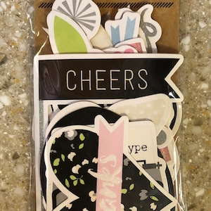 Puede incluir: Un paquete de 24 pegatinas troqueladas con varios diseños. Las pegatinas incluyen una pancarta negra con la palabra "CHEERS", una pancarta rosa con "Thanks" y otras formas decorativas en blanco, negro y verde.
