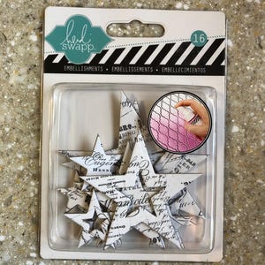 Puede incluir: Un paquete de 16 adornos de estrellas de papel. Las estrellas son blancas con texto y diseños en negro. El paquete está etiquetado como "Heidi Swapp" y "Embellishments".