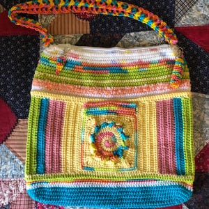 May include: A handmade crochet tote bag with a colorful striped design. The bag features a rainbow-colored strap and a square pocket with a spiral design. The bag's colors include yellow, pink, blue, green, and orange.