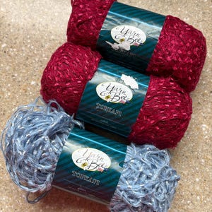May include: Three skeins of Yarn Bee Topkapi yarn. Two skeins are a deep red color, and one is a light blue with silver metallic strands. Each skein has a teal and white label with the brand name and logo.