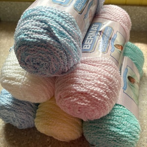 May include: A stack of six skeins of Bernat Baby Coordinates yarn in pastel colors including light blue, white, soft pink, yellow, and mint green. Each skein features a paper label with the brand name and product line printed on the front.