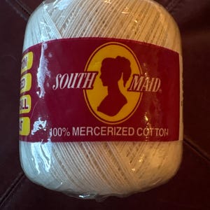May include: A ball of off-white crochet thread wrapped in clear plastic packaging. A red label features the text South Maid, a silhouette of a person, and the words 100% Mercerized Cotton. The thread is wound into a cylindrical shape.