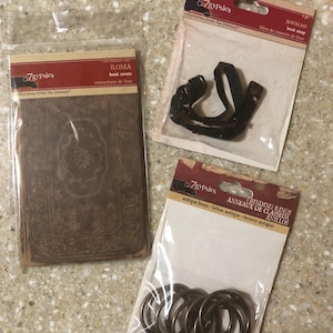 May include: Three craft supplies in clear plastic packaging. One package contains brown book covers, another a black jeweled book strap, and the third, antique brass binding rings. The packaging features the brand name "7gypsies".