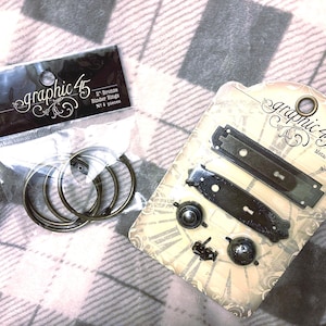 May include: Two packages of Graphic 45 craft supplies. One contains bronze binder rings, and the other includes metal door plates and knobs. The packaging features the Graphic 45 logo and product details.