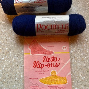 May include: Two navy blue yarn skeins labeled "Rochelle" and "Orlon Sayelle" sit above a pink pattern instruction sheet for "Siesta Slip-ons." The sheet features illustrations of knitted items and text detailing yarn and needle requirements.