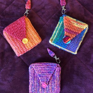 May include: Three handmade crochet coin purses in vibrant colors. One is pink and orange, another is rainbow-colored, and the third is purple and blue. Each purse has a button closure and a small strap with a metal clasp.