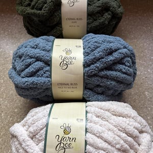 May include: Three skeins of Yarn Bee Eternal Bliss yarn in olive, blue, and ivory. Each skein features a paper label with the brand logo, color name, and net weight of 8 oz. The yarn has a thick, plush, and soft chenille-style texture.
