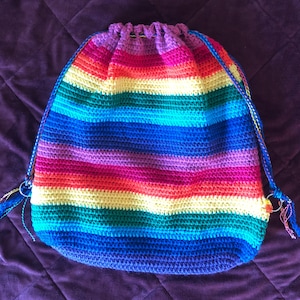 May include: A handmade drawstring backpack in a vibrant rainbow pattern. The bag is crocheted with horizontal stripes of red, orange, yellow, green, blue, and purple. It features blue drawstrings and metal rings for closure.
