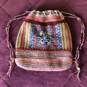 May include: A handmade crochet drawstring backpack in shades of pink, tan, blue, and burgundy. The bag features a central square design with a floral motif and braided drawstring straps. The bag is approximately 12 inches tall.
