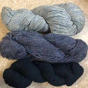 May include: Three skeins of yarn in varying shades of blue and black. The top skein is a light blue with flecks of white, the middle is a darker blue with a speckled appearance, and the bottom is a solid black.