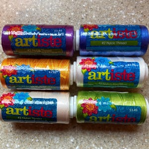 May include: Six spools of Artiste #2 Nylon Thread in various colors: purple, blue, gold, white, and green. Each spool has a label with the brand name and floral design. The thread is wound around a white plastic core.
