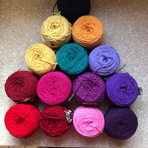 May include: A collection of colorful yarn balls in various shades, including yellow, orange, teal, purple, pink, red, and black. The yarn is arranged in a triangular shape, showcasing the diverse color palette available for crafting projects.