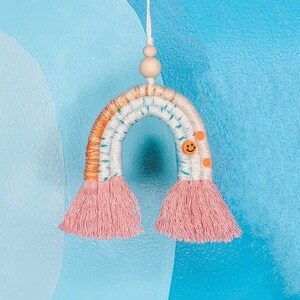 Może przedstawiać: Wiszący makramowy tęczowy dekor ścienny w kolorze różowym i pomarańczowym z uśmiechniętym guzikiem. Tęcza jest zawieszona na drewnianej koralce.