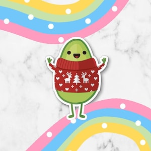 Könnte beinhalten: Eine grüne Avocado, die einen roten Weihnachtspullover mit einem weißen Rentiermuster auf einem weißen Marmorhintergrund mit einem Regenbogen aus Pastellfarben trägt.