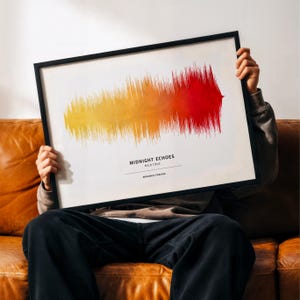 May include: A framed print with a sound wave design in yellow, orange, and red hues. The text reads "MIDNIGHT ECHOES MILO CRUZ MEMORIES FOREVER". The frame is black, and the print is held up by a person.