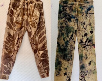 Könnte beinhalten: Zwei handgefertigte Hosen mit einzigartigen Blättermustern. Eine Hose ist braun mit einem Blattmuster, die andere ist beige mit grünen und blauen Blättermustern. Beide haben einen Kordelzugbund und hängen an schwarzen Kleiderbügeln.