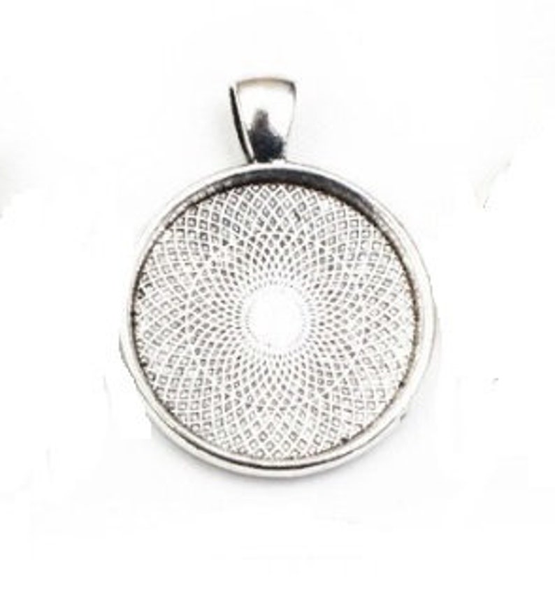 May include: A circular silver-toned metal pendant tray featuring a raised bail at the top. The interior of the pendant base displays a detailed, repeating geometric pattern of small, interlocking diamond shapes radiating from the center.