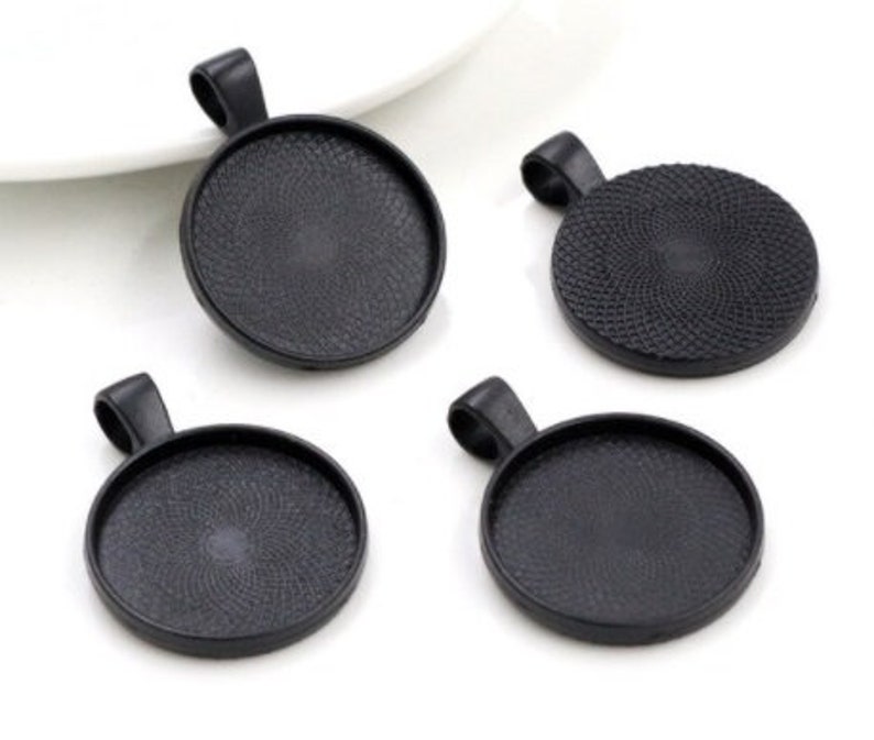 May include: Four circular black pendant trays with a textured interior base and a top loop for hanging. These jewelry findings feature a matte black finish and a recessed center designed for setting cabochons or other decorative elements.