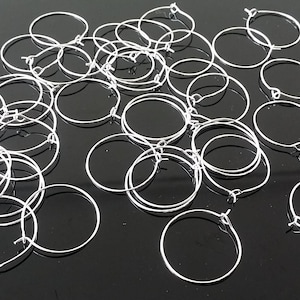 May include: A collection of small, silver-coloured hoop earring findings. The open hoops are made of thin wire and are scattered across a dark, reflective surface. These findings are used for jewellery making.
